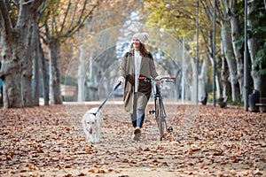 Beautiful young girl riding a bike while walking her dog in the park in autumn