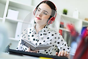 Beautiful young girl sits in headphones at desk in office, holds a notebook in her hand and prints on the keyboard