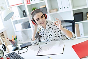 A beautiful young girl sits in headphones and with a microphone at the desk in the office and shows a sign of okay.