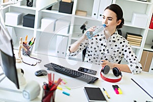 A beautiful young girl sits at a table in the office and drinks water from a bottle.