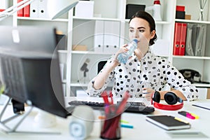 A beautiful young girl sits at a table in the office and drinks water from a bottle.