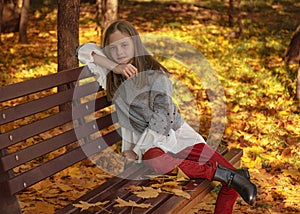 Beautiful young girl sitting on a brown wooden park bench in autumn. Lady thought and dreams. Autumn leaf fall in the city.
