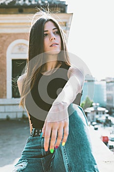 Beautiful young girl sitting in the city. Urban outfit and style, jeans and black top. Colored nail polish. Relaxed, bright light