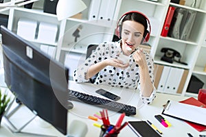 Beautiful young girl sitting in headphones at desk in office, eating yogurt and looking at monitor.