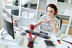 Beautiful young girl sitting in headphones at desk in office, eating yogurt and looking at monitor.