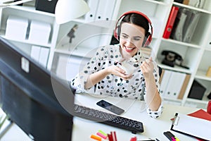 Beautiful young girl sitting in headphones at desk in office, eating yogurt and looking at monitor.