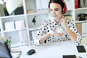 Beautiful young girl sitting in headphones at desk in office, eating yogurt and looking at monitor.