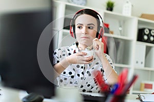 Beautiful young girl sitting in headphones at desk in office, eating yogurt and looking at monitor.