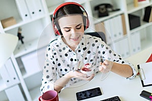 Beautiful young girl sitting in headphones at desk in office eating yogurt with red filling.