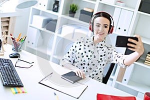 Beautiful young girl sitting in headphones at desk in office and taking pictures of herself on the phone. A photograph