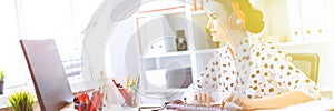 Beautiful young girl sitting in headphones at desk in office and typing on keyboard.