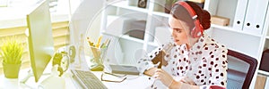 Beautiful young girl is sitting in headphones and with a microphone at the desk in the office.