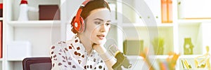 Beautiful young girl sitting in headphones and with microphone at table in office and talking.