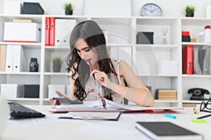 Beautiful young girl sitting at an office desk holding a pen in her mouth and looking into the calculator.