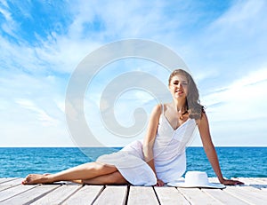 Beautiful, young girl sitting on a pier in a white dress. Summer, vacation and traveling concept.