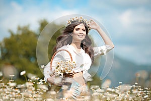 Beautiful young girl smiling with basket of flowers over chamomile field. Carefree happy brunette woman with healthy wavy hair photo