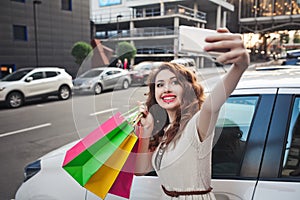 Beautiful young girl stands near a white car, makes a shopping, selfie