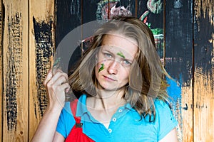 Beautiful and young girl stands on a wooden wall background which is painted with black paint. The girl`s face is stained with gre