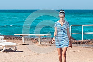 Beautiful and young girl in a striped blue dress walks against the backdrop of the blue sea with a small stripe the sky