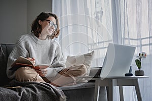 Beautiful young girl student studying at home online while sitting in a warm sweater on the couch in front of a laptop making