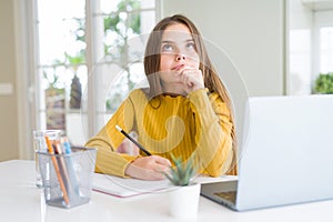 Beautiful young girl studying using computer laptop and writing on notebook serious face thinking about question, very confused