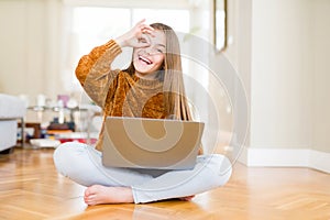 Beautiful young girl studying using laptop sitting on the floor at home with happy face smiling doing ok sign with hand on eye