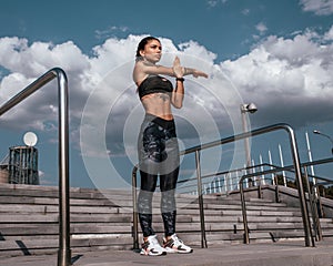 Beautiful young girl in tattoos, summer city, stretching muscles of hands before training sportswear leggings and bra