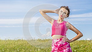 Young beautiful girl in pink dress stands on green lawn against blue cloudy summer sky.