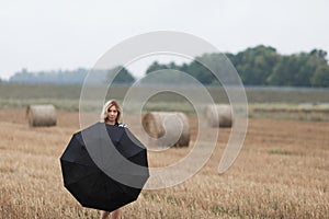 A beautiful young girl with an umbrella is standing in a field near a haystack.