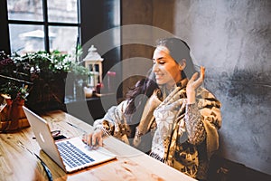 Beautiful young girl uses laptop technology, types text looking at the monitor in a cafe by the window at wooden table