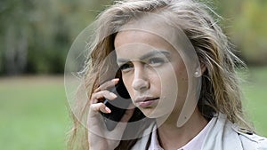 Beautiful young girl using her cell phone, outdoor. Portrait of relaxed young lady in a summer park talking on phone