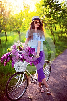 Beautiful young girl with vintage bicycle and flowers on city background in the sunlight outdoor.