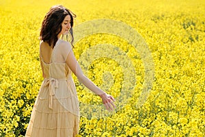 Beautiful young girl walking in the yellow canola field, touching flowers with her hand. Model dressed in beige dress relaxing