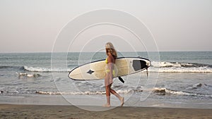 A beautiful young girl walks along a sandy beach with a surfboard