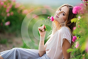 Beautiful young girl is wearing casual clothes having rest in a garden with pink blossom roses