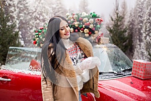 Beautiful young girl is wearing winter clothes holding sparklers near red car with decorated xmas tree on the roof