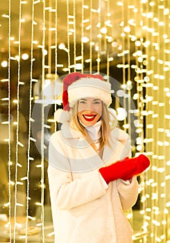 A beautiful, young girl in a white fur coat and Santa hat, standing near the glowing lights, smiling and laughing