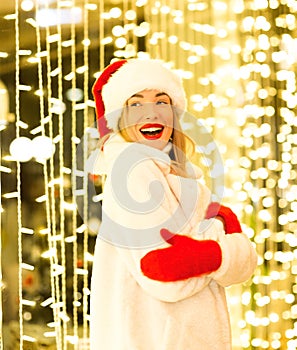 A beautiful, young girl in a white fur coat and Santa hat, standing near the glowing lights, smiling and laughing