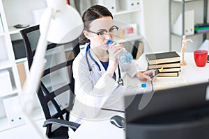 A beautiful young girl in a white robe sits at a computer desk, drinks water and looks at the monitor.