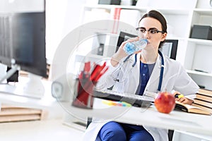 A beautiful young girl in a white robe sits at a computer desk, drinks water and looks at the monitor.