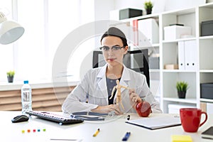 A beautiful young girl in a white robe is sitting at a table and holding a wooden figure of a man in her hands.