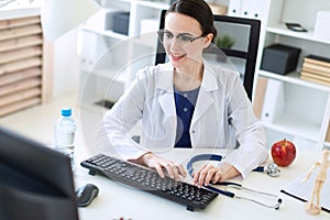 A beautiful young girl in a white robe is sitting at the table and typing on the keyboard.