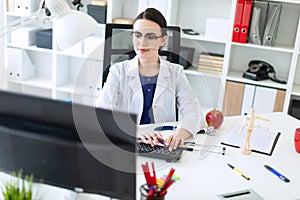 A beautiful young girl in a white robe is sitting at the table and typing on the keyboard.