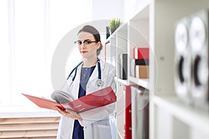 A beautiful young girl in a white robe is standing near the shelter and flips through a red folder with documents.