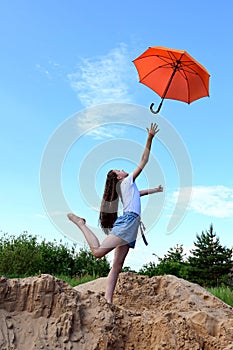 beautiful young girl in white T-shirt and denim shorts throws up an orange umbrella. Sunny summer day outdoors on blue sky