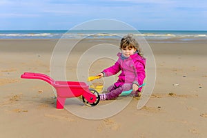 Beautiful young girl in winter clothes playing on the beach with her wheelbarrow and plastic toys