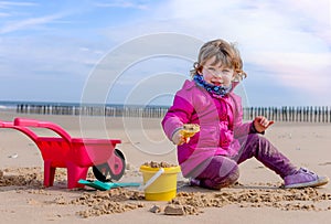 Beautiful young girl in winter clothes playing on the beach with her wheelbarrow and plastic toys