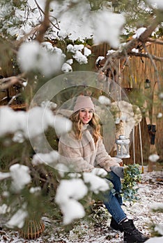 A beautiful young girl in winter clothes sits under a snowy pine tree