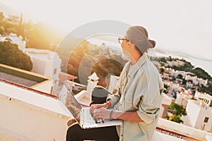 Beautiful young girl woman in eyeglasses sitting with a laptop on her balcony at sunset with a view of the city, remote work from