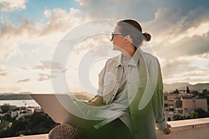 Beautiful young girl woman in eyeglasses sitting with a laptop on her balcony at sunset with a view of the city, remote work from